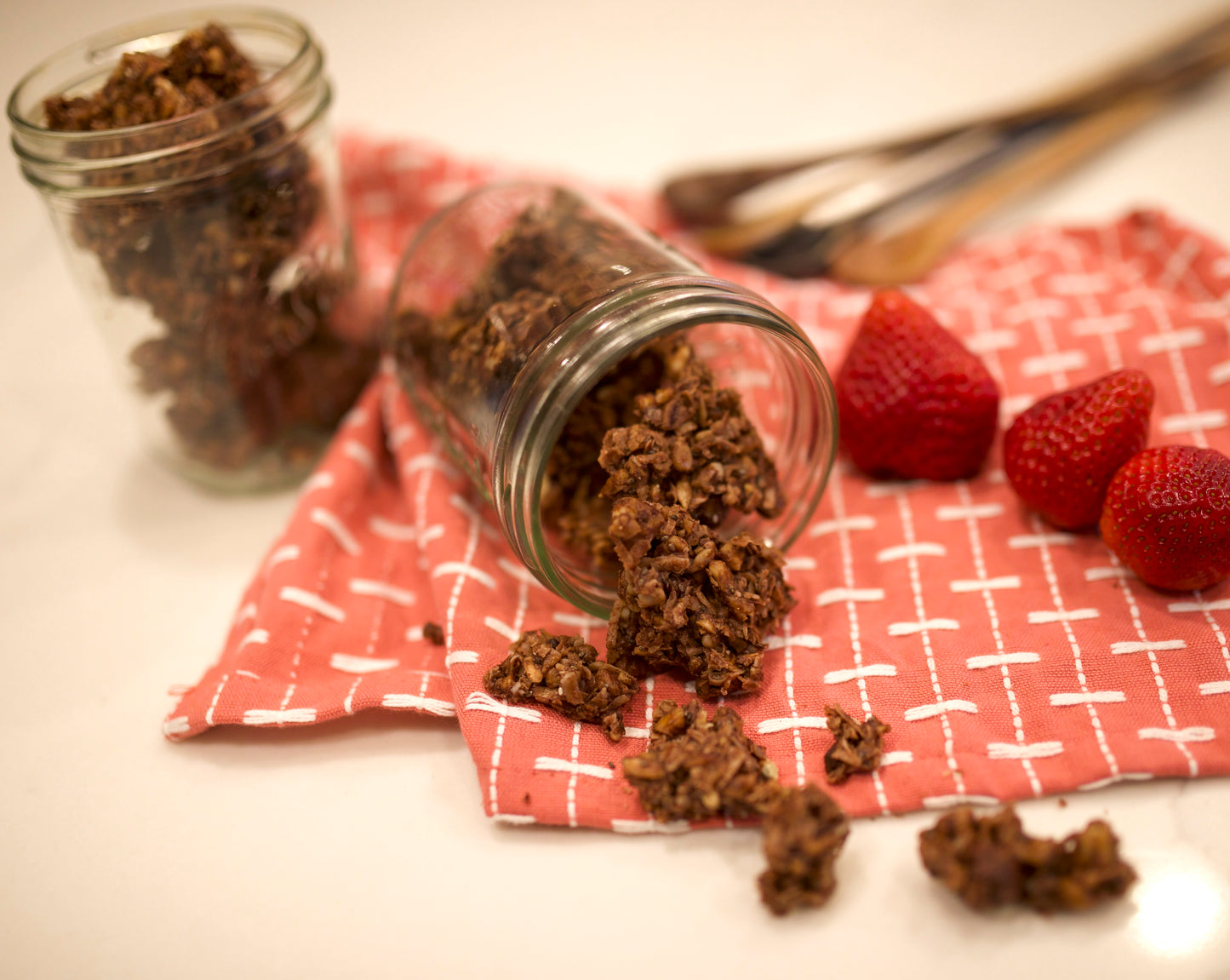 Glass jars with chocolate-covered strawberries on a red checkered cloth with strawberries nearby.