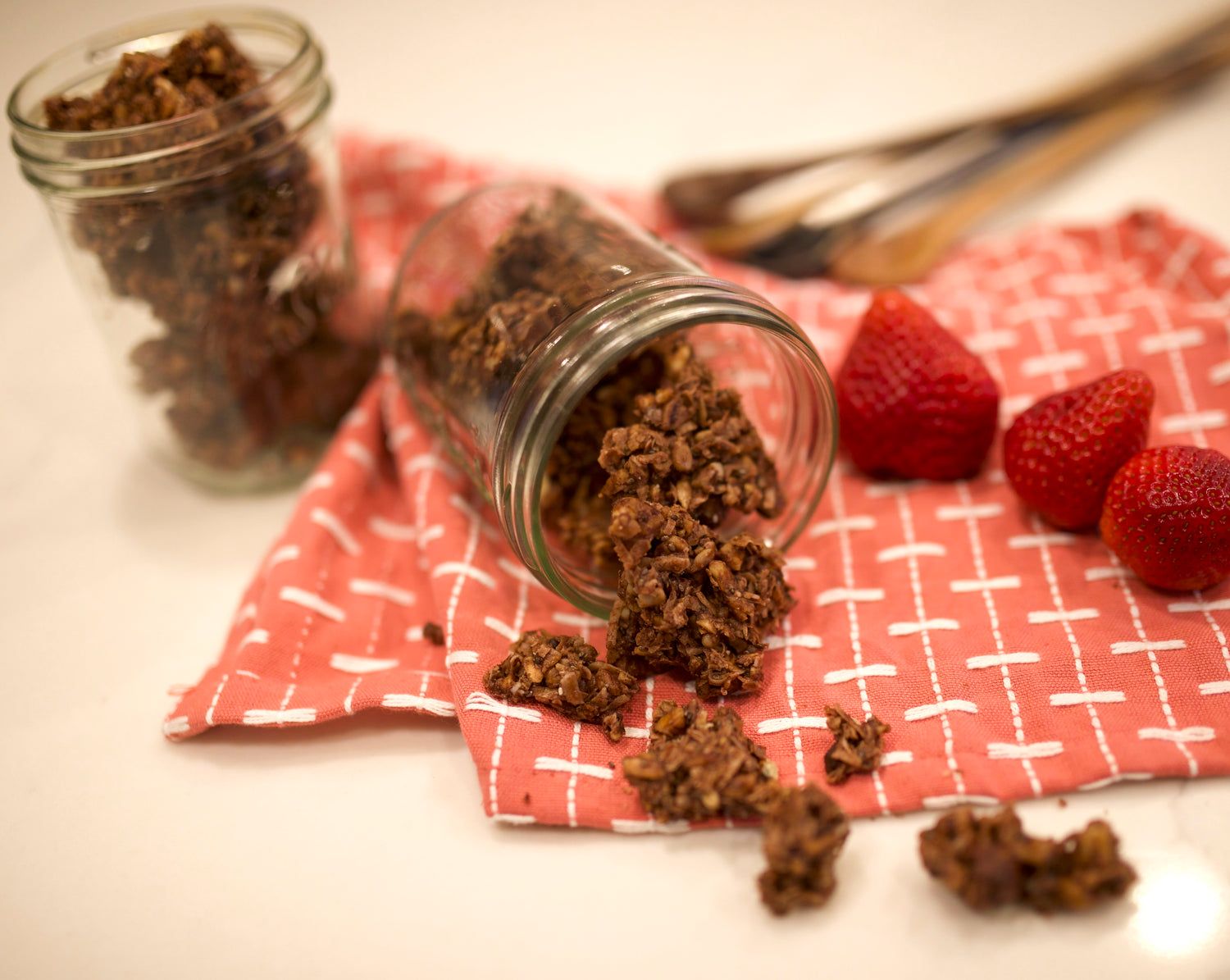Glass jars with chocolate-covered strawberries on a red checkered cloth with strawberries nearby.